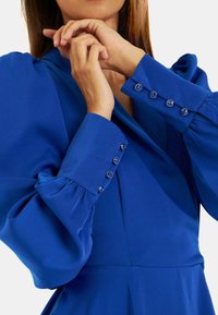 Blaues Kleid mit puffierten Ärmeln und V-Ausschnitt, aus glattem Stoff gefertigt. Knopfdetails an den Bündchen fügen ein dekoratives Element hinzu.