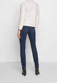 Světlá béžová péřová bunda s prošívaným designem a vysokým límcem. Kombinována s tmavě modrými úzkými džínami a černými kotníkovými botami na podpatku.