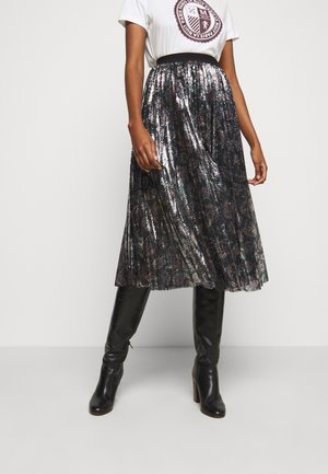 Vrouw draagt een wit grafisch t-shirt, zwarte en zilveren metallic geplooide midi rok, en zwarte kniehoge laarzen met hakken tegen een effen achtergrond.