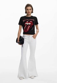 Mustast puuvillast T-särk, millel on punane Rolling Stonesi logo, koos valgete lehvidega teksadega. Modell hoiab väikest musta kotti ja kannab valgeid kingi.