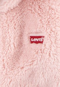 Duslus rožinis fliso audinys su švelniu tekstūra. Yra raudona Levi's logotipo aplikacija, siuvinėta į medžiagą.