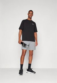Svart t-skjorte med korte ermer og hvit logo, kombinert med grå shorts. Modellen holder en svart vannflaske og har på seg svarte joggesko.