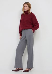 Frau steht an einer weißen Wand und trägt einen strukturierten burgunderfarbenen Rollkragenpullover, weite graue Hosen und burgunderfarbene Kitten-Heel-Schuhe.