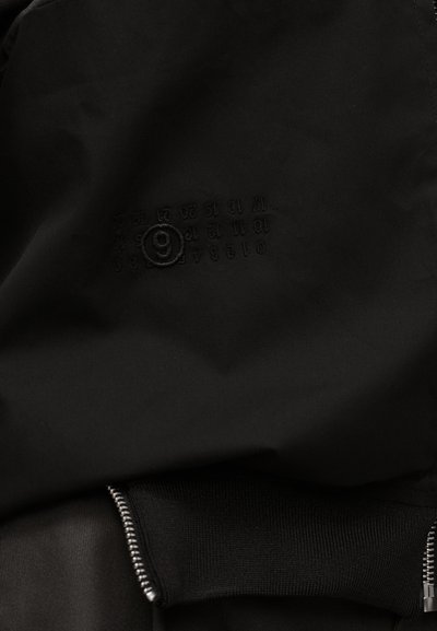 Μαύρο ύφασμα με ραμμένους αριθμούς διευθετημένους σε σειρές, ο αριθμός 9 είναι κυκλωμένος, και εμφανίζεται ένα ασημί φερμουάρ με μαύρο ύφασμα με ρίγες.