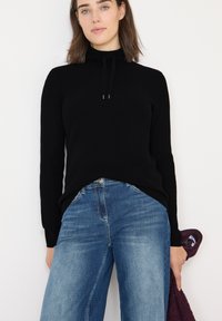 Maglione nero a costine con colletto alto e coulisse. Abbinato a jeans a gamba ampia blu. Mano che tiene un oggetto bordeaux. Design semplice e moderno.