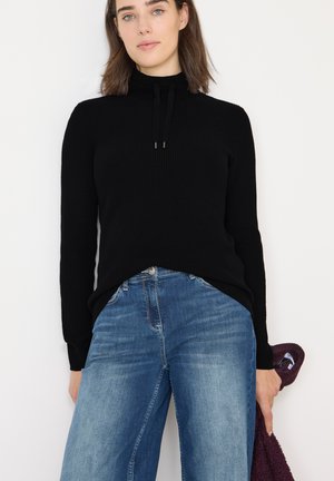 Pull noir côtelé avec un col montant et un cordon de serrage. Associé à un jean bleu à jambes larges. Main tenant un objet bordeaux. Design simple et moderne.