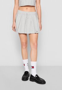 Femme portant une mini-jupe plissée gris clair avec cordon, des chaussettes blanches à rayures rouges et bleu marine, et des mocassins noirs, debout devant un fond uni.