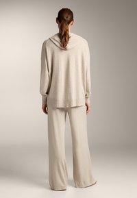 Svetlé béžové loungewear oblečenie pozostávajúce z mikiny s kapucňou a dlhými rukávmi a nohavíc so širokými nohavicami. Mäkkučký materiál s voľným strihom a rebrovanými manžetami.