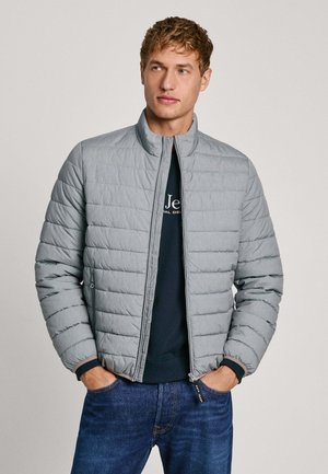 Veste grise rembourrée avec col montant et fermeture éclair, dotée de coutures horizontales et de poignets côtelés, portée par-dessus une chemise sombre et un jean.