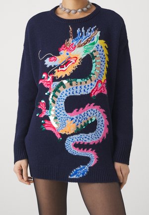 Jersey azul marino con diseño de dragón bordado y con lentejuelas de colores, combinado con medias negras transparentes y un collar floral plateado.