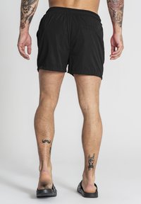 Bañador negro con cintura elástica, de corta longitud y dos bolsillos laterales. Tela texturizada con detalles de costura visibles, combinado con chanclas.