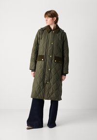 Olijfgroene gewatteerde jas met een rits- en knoopsluiting, corduroy accenten op de zakken en een met bont gevoerde kraag. Draag het over donkere wijde jeans.