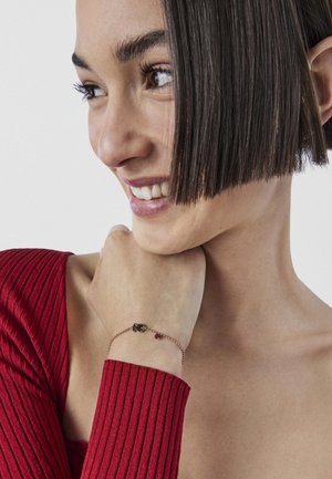 Bracciale a catena in tonalità oro con un piccolo ciondolo scuro e una perlina rossa. Indossato sul polso, con una cima rossa a trama visibile.