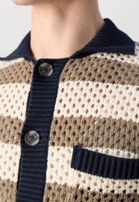 Gros plan sur un cardigan en maille beige et blanc à rayures, avec col côtelé bleu marine, patte de boutonnage, deux boutons, et détail de poche.