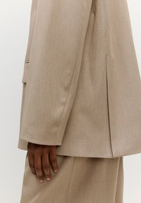 Beige op maat gemaakte blazer van textuurstof, met een enkel ventilatieopening aan de achterkant en een zak aan de voorkant aan de linkerkant.