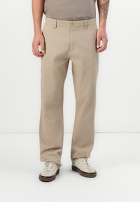 Man wearing beige straight-leg pants, light beige sneakers, and a taupe t-shirt against a white background.