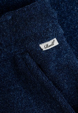 Dunkelblaues Frottiergewebe mit einer strukturierten Oberfläche. Verfügt über ein kleines weißes Etikett mit der Aufschrift "Reell", das darauf gestickt ist.