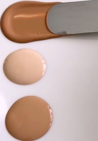 Tre flytende kosmetiske nyanser: lys beige, mellom beige og dyp tan, påført i sirkulære flekker på en hvit overflate med en spatel.