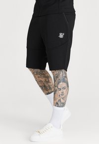 Mustat urheilushortsit ommelluilla yksityiskohdilla, sivutaskut ja valkoinen logo. Mallilla on valkoiset kengät kultaisilla yksityiskohdilla ja valkoiset sukat.