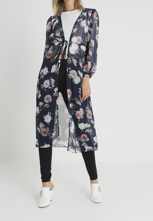 Frau trägt eine marineblaue, florale, transparente lange Strickjacke über einem weißen Oberteil, schwarzen Hosen und weißen Schuhen mit Absatz, steht vor einem schlichten Hintergrund.