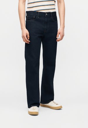 Personne portant un jean noir droit, des baskets blanc cassé avec des semelles en gomme, et une chemise blanche avec des rayures bleues et orange.