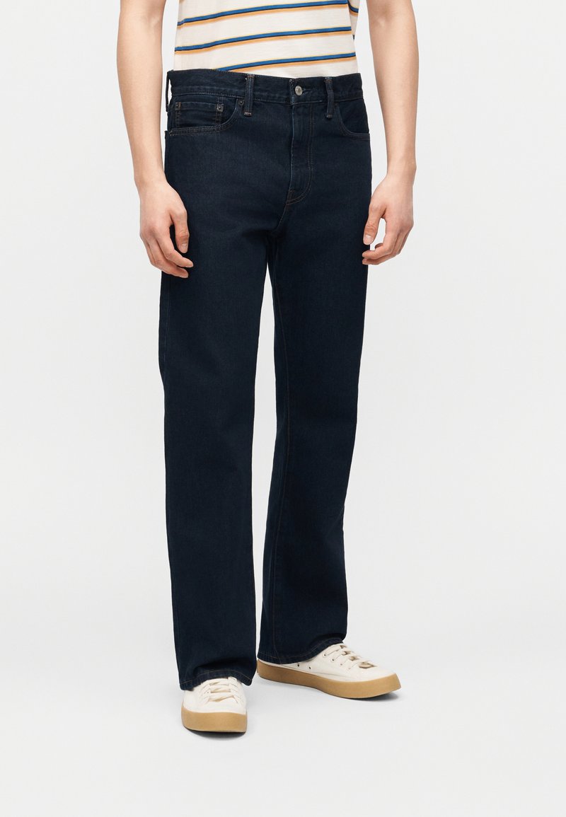 Άτομο που φοράει ίσια μαύρα τζιν, εκρού αθλητικά παπούτσια με λαστιχένιες σόλες και λευκό πουκάμισο με μπλε και πορτοκαλί ρίγες.