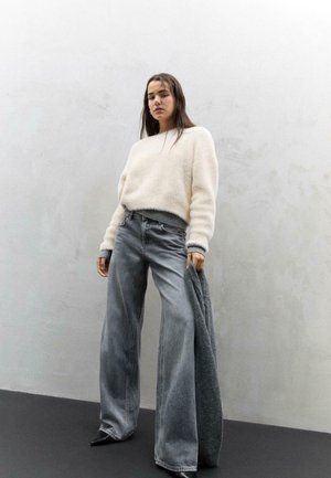 Cremefarbener, flauschiger Pullover mit gerippten Ärmelbündchen, kombiniert mit weit geschnittenen grauen Jeans. Das Model hält eine dunkelgraue Strickjacke und steht auf einer schwarzen Oberfläche.