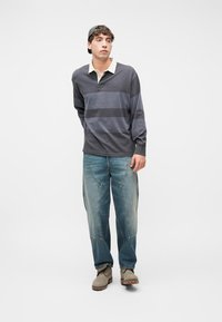 Νέος άνδρας που φοράει σκούρο ριγέ μακρυμάνικο πόλο, ξεβαμμένο χαλαρό τζιν, μπεζ μπότες και μαύρο καπέλο προς τα πίσω, στέκεται σε λευκό φόντο.