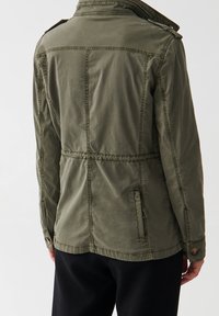 Chaqueta ligera verde oliva con cuello alzado, bolsillos laterales y cintura elástica. Presenta detalles de costura y acentos de botones.