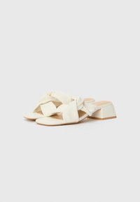 Sandales à talon carré de couleur crème avec un design de bride nouée et une texture lisse, offrant une finition mate et une semelle intérieure beige subtile.