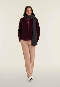 Giacca in pile color bordeaux con zip, t-shirt color malva, pantaloni della tuta beige e una sciarpa grigia scura. Sneakers bianche completano il look.