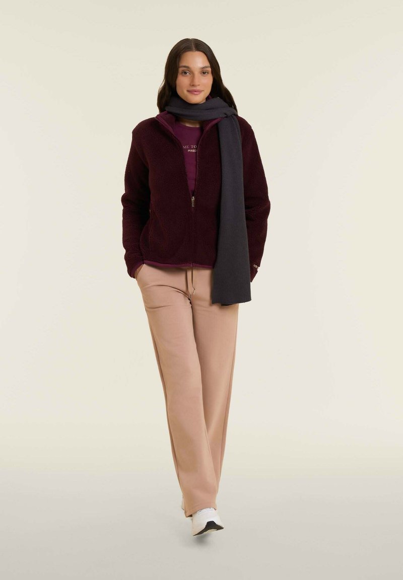 Giacca in pile color bordeaux con zip, t-shirt color malva, pantaloni della tuta beige e una sciarpa grigia scura. Sneakers bianche completano il look.