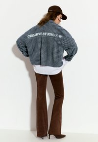 Giacca a scacchi blu e bianca tagliata, con il testo "CREATIVE STUDIO" sulla schiena, abbinata a pantaloni svasati marroni e tacchi scuri.