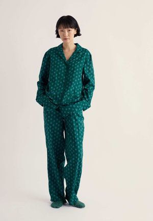 Femme debout les mains dans les poches, portant un ensemble pyjama à manches longues vert à motifs avec des chaussettes assorties sur un fond neutre.