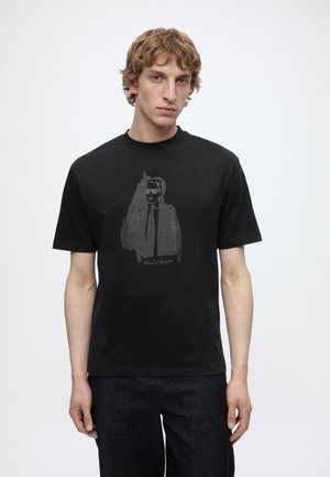 Junger Mann mit hellem lockigem Haar, der ein schwarzes T-Shirt mit einem dunklen Motiv eines Mannes im Anzug und mit Sonnenbrille trägt.