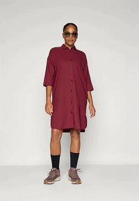 Vestido burdeos de botones con mangas cortas, que cae por encima de la rodilla. Llevado con calcetines oscuros y zapatillas marrones. Textura de tela suave.