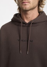 Haut à capuche marron avec un cordon de serrage, présentant un logo "VOLCOM" brodé en noir sur le devant. Matière douce et coupe décontractée.