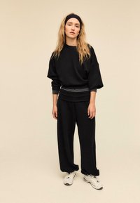 Schwarzer übergroßer Pullover mit gestreiften Bündchen, kombiniert mit lockeren schwarzen Hosen. Das Modell trägt ein Stirnband und weiße Sneaker. Einfache, weiche Texturen.