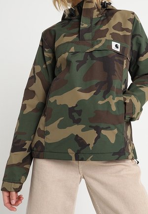 Personne portant une veste camouflage verte et marron avec une poche frontale et un pantalon beige, debout avec une main dans la poche.