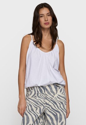 Donna con lunghi capelli scuri indossa una maglietta bianca senza maniche larga e pantaloni a vita alta con righe zebra bianche e nere, in piedi davanti a uno sfondo semplice.