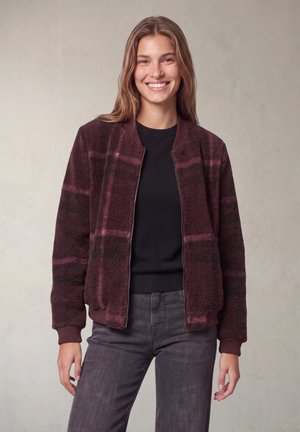 Jeune femme souriante portant une veste polaire à carreaux bordeaux et rose, un pull noir et un jean gris foncé sur un fond neutre.