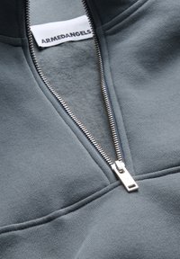 Sudadera gris con cremallera, que presenta una textura suave, cremallera plateada y una etiqueta que dice "ARMEDANGELS" situada cerca del escote.