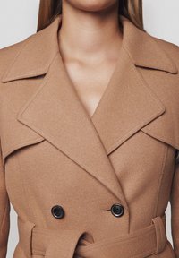 Dwurzędowy płaszcz z miękkiej wełny w odcieniu camel, z szerokim kołnierzem, czarnymi guzikami i paskiem w talii, tworzącym zarys sylwetki.