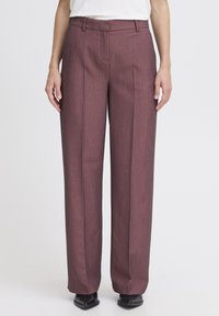 Pantaloni larghi color marrone in tessuto texturizzato, con fronte piatto, passanti per cintura e dettagli di piega; abbinati a scarpe nere.