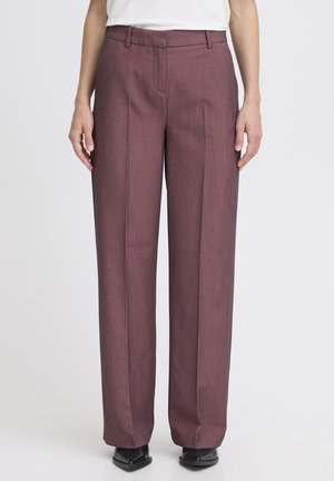 Pantaloni larghi color marrone in tessuto texturizzato, con fronte piatto, passanti per cintura e dettagli di piega; abbinati a scarpe nere.
