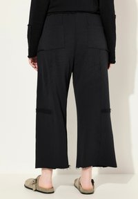Pantaloni neri a gamba larga realizzati in morbido tessuto con orlo sfrangiato, dotati di due tasche posteriori e un leggero effetto usurato sui lati.