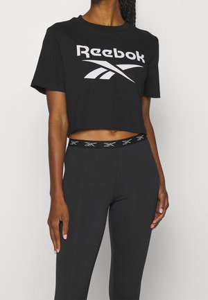 Schwarzes, kurzes Reebok T-Shirt mit weißem Logo, getragen mit schwarzen Leggings mit sich wiederholendem Reebok-Logo am Bund.