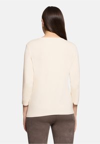 Cremefarbenes Langarmshirt mit rundem Ausschnitt und glatter Textur, kombiniert mit grauer Hose, von hinten gezeigt.