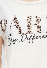 T-shirt blanc à manches courtes arborant le mot "PARIS" en lettres épaisses avec un motif léopard, et "Stay Different" en cursive en dessous.