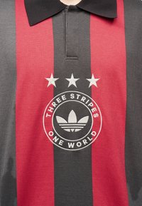 Πόλο πουκάμισο με κόκκινες και σκούρες γκρι κάθετες ρίγες, τρία λευκά αστέρια πάνω από έναν κυκλικό λογότυπο που γράφει "Three Stripes One World" και ένα σχέδιο τριφυλλιού.